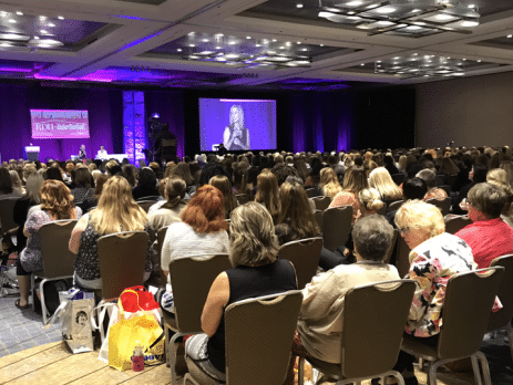 View from behind an audience sitting in a conference room watching a woman speak on stage