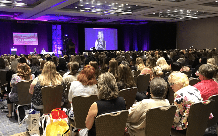 View from behind an audience sitting in a conference room watching a woman speak on stage