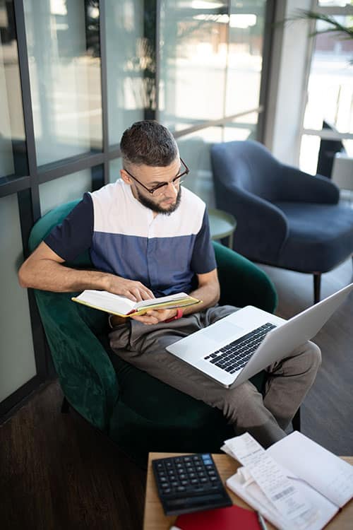 A man references a book while working on his laptop